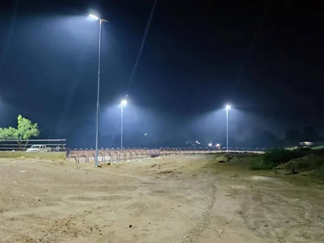 A nighttime view of a sandy outdoor area with three tall streetlights shining against a dark sky.