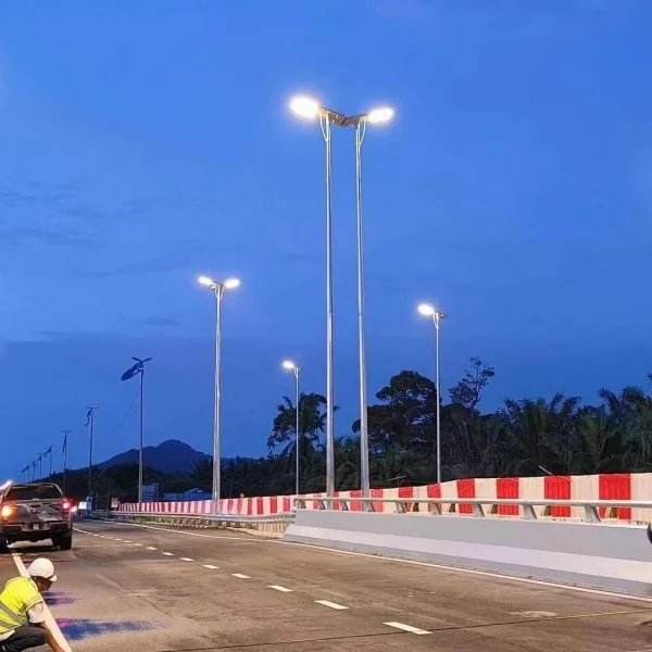 A newly constructed road at dusk features tall dual-headed solar street lights, red and white barriers, and a worker in the foreground.
